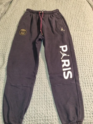 Jordan PSG svarta mjukisbyxor - Svarta mjukisbyxor från Jordan  PSG med guldfärgade detaljer och loggor. Byxorna har resår i midjan med lila snörning, fickor på sidorna och trycket 'PARIS' längs ena benet. Fast pris.