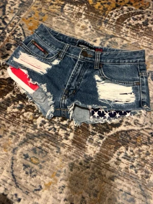 Blå denimshorts med USA design  - Säljer ett par blå jeansshorts från Maro Cat med slitna detaljer och råa kanter. Shortsen har framfickor med vita stjärnor på marinblå botten och röda ränder på vit botten som syns genom de slitna partierna. Klassisk femficksmodell och knappgylf. Storleken är W26.