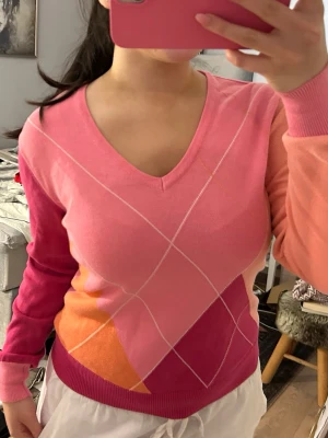 Färgglad v-ringad tröja i rosa och orange - Säljer en snygg v-ringad tröja med argylemönster i olika nyanser av rosa, orange och lite vitt. Tröjan har långa ärmar och är i mjukt, tunt material. Perfekt för dig som gillar färg och vill sticka ut lite extra. Stor inget märke och storleken är uppskattningsvis en M