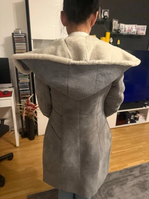 Grå lång pälsjacka/kappa med huva - Säljer en lång och snygg grå pälsjacka/ kappa med stor fluffig huva och mjukt vitt foder. Jackan har dragkedja framtill, två fickor och quiltade detaljer vid ärmslut och midja. Perfekt för kalla dagar när du vill vara både varm och stilren. Ordinarie pris 3000kr