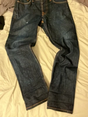 Mörkblå jeans från Nudie Jeans, 31/30 - Säljer ett par mörkblå jeans från Nudie Jeans i klassisk straight fit. Jeansen har orangea sömmar och snygga detaljer på bakfickorna. Tillverkade i slitstarkt jeansmaterial av bomull. Perfekta för dig som gillar en tidlös och clean look.