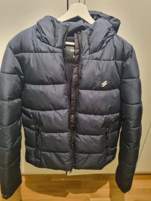 Mörkblå pufferjacka från Superdry - Säljer en mörkblå pufferjacka från Superdry med huva och dragkedja. Jackan har två fickor med dragkedja och diskret logga på bröstet. 