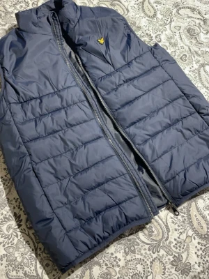 Mörkblå dunväst från Lyle & Scott - Säljer en mörkblå dunväst från Lyle & Scott med gul logga på bröstet. Västen har hög krage, quiltad design och dragkedja framtill. Perfekt att slänga över en hoodie eller tröja för extra värme. Enkel och stilren look. Den är igentligen storlek xl i junior men sitter som S