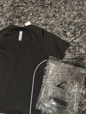 Svart Gymshark t-shirt herr L - Svart Gymshark t-shirt i storlek L men kan passa M oxå, med vita detaljer längs sidorna och logga på bröstet. Tillverkad i ett stretchigt och lätt material som passar perfekt för träning. Snygg och enkel design med rund hals och kort ärm.