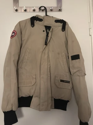 Beige bomberjacka från Canada Goose - Säljer en beige bomberjacka från Canada Goose med svart foder och detaljer. Jackan har huva, ribbade muddar, flera fickor och dragkedja framtill. Klassisk patch på ärmen och snygg, sportig look. Perfekt för kyliga dagar och riktigt stilren. Fläckar men ska tvätta jackan senast imorgon 