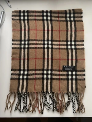 Rutig halsduk från Burberry i kashmir - Klassisk halsduk från Burberry i 100% kashmir med det ikoniska rutiga mönstret i beige, svart, vitt och rött. Halsduken har fransar i ändarna och är tillverkad i England. Perfekt accessoar för att lyfta din outfit.