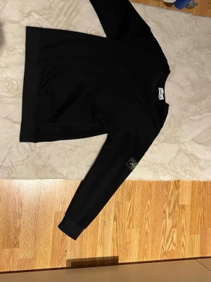 Svart sweatshirt från Stone Island - Svart sweatshirt från Stone Island med klassisk rund hals och ribbade muddar. Tröjan har den ikoniska Stone Island-loggan på vänster ärm. Tillverkad i mjuk bomull och har en clean, stilren look som passar perfekt till streetwear. Säljes för den inte passade mig, storlek L fast liten i storlek, pasar S/M.