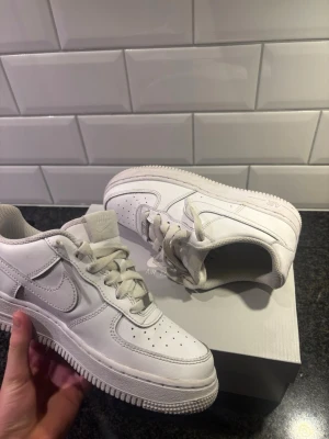 Nike Air Force 1 vita sneakers - Klassiska Nike Air Force 1 sneakers i helvitt skinn med perforerad tå, rund tå och snörning. Snygg och tidlös modell med låg profil och vit sula. Perfekta till streetwear och passar till det mesta. Kommer i originalkartong.