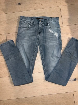 Ljusblå skinny jeans från Nudie - Säljer ett par ljusblå skinny jeans från Nudie med slitningar och tvättade detaljer på framsidan. Klassisk femficksmodell med snygga sömmar på bakfickorna. Jeansen är i stretchigt denim och har normal passform i midjan.25/32