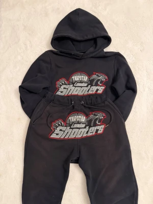 Trapstar dress - Trapstar svart hoodie och byxor med stort broderat 'London Shooters'-motiv i grått, vitt och rött. Setet har huva, dragsko i midjan och dragkedjor vid bensluten. Materialet är mjukt och skönt, perfekt för chill eller streetwear. Passar Xs och S