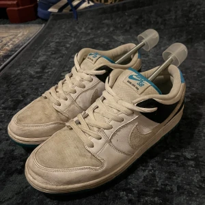 Nike SB Dunk Low Pro vit/blå - Nike SB Dunk Low Pro sneakers i vitt och svart med blå detaljer på plösen och hälen. Skorna har snörning, rund tå och en blågrön yttersula. Ovandelen är i mocka och skinn, vilket ger en schysst kontrast mellan materialen. Perfekta för dig som gillar klassiska skatevibes. Storlek 44