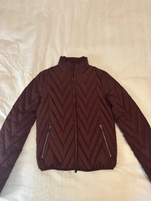 Mörkröd quiltad pufferjacka Selected Femme - Snygg mörkröd pufferjacka från Selected Femme i storlek 36. Jackan har quiltat mönster i zigzag, hög krage och två fickor med dragkedja. Tillverkad i polyester och perfekt för kyligare dagar. Stängs med dragkedja framtill.