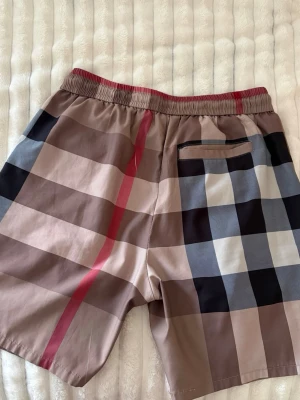 Rutig Burberry shorts med snörning - Säljer ett par klassiska Burberry shorts i bomull med det ikoniska rutiga mönstret i beige, svart, vit och röd. Shortsen har elastisk midja med svart snörning och fickor både fram och bak. Perfekta för sommaren och riktigt snygga till en enkel t-shirt.