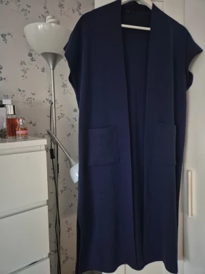 Mörkblå lång stickad väst - Säljer en mörkblå lång stickad väst med två stora fickor framtill. Västen är ärmlös, har rak passform och är tillverkad i ett mjukt material. Perfekt att slänga över en tröja eller skjorta för en chill look.