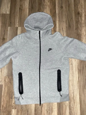 Grå Nike Tracksuit - Säljer ett par grå mjukisbyxor och Hoodie från Nike med svart logga på vänster sida. Byxorna har elastisk midja med svartvit snörning, muddar vid bensluten och praktiska fickor med dragkedja. Perfekta för chill eller träning. Ny pris runt 3000kr