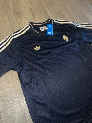Adidas performance shirt - Adidas performance Juventus jersey, med tre vita ränder på ärmarna och en guldfärgad logotyp på bröstet. Tröjan har ett mönster av vertikala ränder och en rund halsringning med guldfärgade detaljer.  Nypris: 1199:-