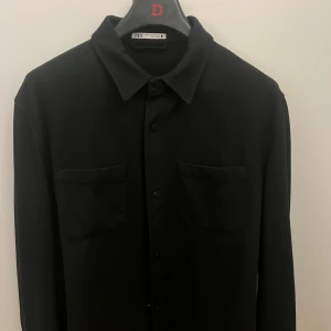 Svart overshirt från Zara - Stilren svart overshirt från Zara med klassisk krage, två bröstfickor och knäppning framtill. Skjortan har lång ärm och en rak, något loose passform. Perfekt att slänga över en t-shirt för en clean look.