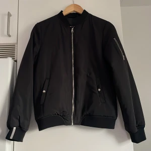 Svart bomberjacka från D-WAY - Svart bomberjacka från D-WAY med silvriga dragkedjor, ribbade muddar med reflex och fickor med tryckknappar. Jackan har en klassisk bomberkrage och en extra ficka med dragkedja på ärmen. Materialet är slätt och jackan har en clean, stilren look. Köpt ny och aldrig använd. 