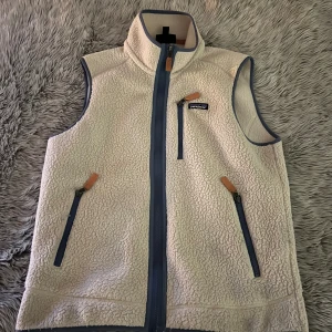 Beige fleeceväst från Patagonia - Mysig beige fleeceväst från Patagonia med hög krage och blå detaljer. Västen har tre fickor med dragkedja och snygga läderdetaljer på dragkedjorna. Perfekt lager-på-lager-plagg för kyliga dagar.