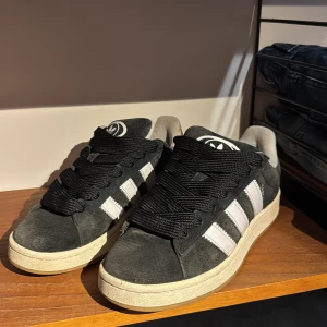 Adidas Campus svarta sneakers i mocka - Svarta Adidas Campus sneakers i mjuk mocka med klassiska vita ränder på sidorna och vit Adidas-logga på hälen. Skorna har breda svarta snören, vit sula och gummiyttersula med fiskbensmönster. Perfekta för en streetwear-look.