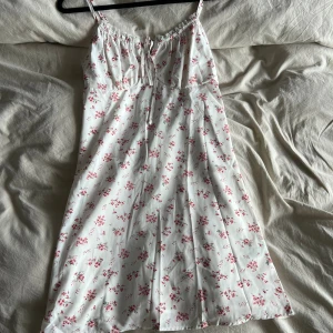 Vit blommig sommarklänning med smala axelband - Supersöt vit klänning med små rosa blommor och smala axelband. Klänningen har en lätt utställd form, knytning framtill och är perfekt för varma dagar. Tunt och luftigt material som ger en romantisk vibe.