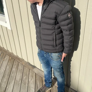 Svart Colmar pufferjacka med huva - Säljer en svart pufferjacka från Colmar med huva och dragkedja framtill. Jackan har ett stilrent, quiltat mönster och logotyp på ärmen. Perfekt för kyliga dagar och ger en clean look till jeans och sneakers.