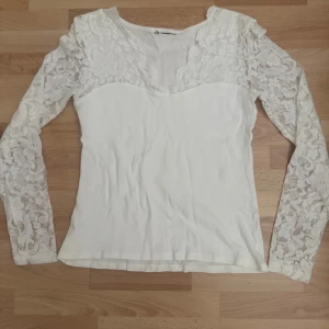 Vit spetsblus med lång ärm - Supersöt vit blus med långa ärmar i blommig spets och v-ringad halsringning. Toppen har en figurnära passform och är tillverkad i mjukt material med spetsdetaljer över axlar och ärmar. Perfekt för dig som gillar romantisk och feminin stil. Storlek lapp avklippt.