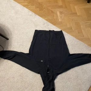 Svart hoodie från Under Armour - Svart hoodie från Under Armour med dragkedja framtill och två stora fickor på magen. Tröjan har huva och långa ärmar, perfekt för en sportig och avslappnad stil. Klassisk logga på ena fickan och mjukt material som känns skönt mot huden.