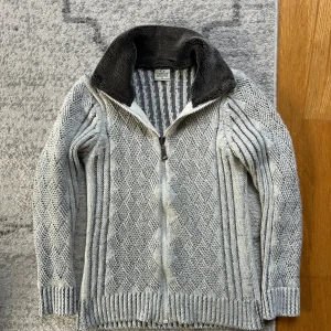 Fodrad Cardigan - Passar som en M, Snygg grå stickad kofta med dragkedja framtill och mysig mörkgrå fleecefodrad krage. Koftan har ett rutigt stickat mönster på framsidan och ärmarna, samt randigt mönster på baksidan. Perfekt för kyliga dagar.