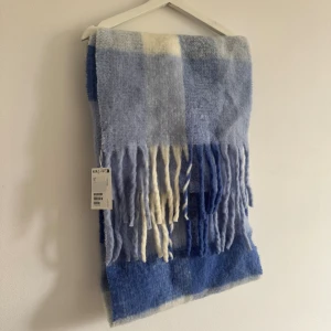 Helt ny rutig ullhalsduk från & Other Stories - Mysig och fluffig halsduk från & Other Stories i rutigt mönster med olika nyanser av blått och inslag av vitt. Gjord av 83% ansvarsfull ull, med långa fransar i ändarna. Perfekt för att hålla värmen och ge din outfit en cool touch. Helt ny med prislapp kvar. Nypris 1090kr