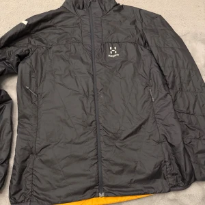 Svart vindjacka från Haglöfs - Helt ny Svart vindjacka från Haglöfs med hel dragkedja framtill och två sidofickor med dragkedja. Jackan har en sportig look, hög krage och diskret logga på bröstet. Insidan har en orange detalj som syns vid nederkanten. Perfekt för uteliv och vardag.