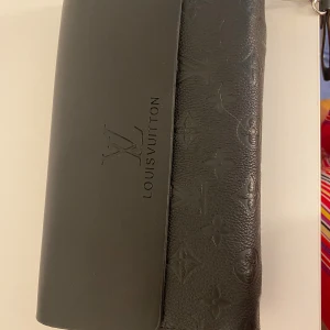 Svart kuvertväska i skinn - Snygg svart kuvertväska med klassiskt monogrammönster i präglat skinn. Väskan har två stora fack med silverfärgade dragkedjor och en avtagbar handledsrem. Perfekt storlek för det viktigaste och stilren design med logga på framsidan. Hel ny