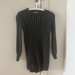 Glittrig svart ribbad mini klänning - Snygg svart ribbad kort klänning med glittriga inslag och långärmad design. Den har ett randigt mönster och sitter figurnära med rund hals. Perfekt för dig som vill sticka ut med lite extra glitter och stil.
