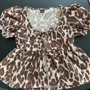Leopardmönstrad blus från Gina Tricot - Säljer en söt blus från Gina Tricot i storlek XS med leopardmönster i bruna och beige toner. Blusen har puffiga korta ärmar, två rosetter framtill och en volang nedtill. Materialet är lätt och ribbat, perfekt för att sticka ut med coola detaljer.