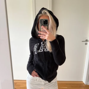 Svart GAP hoodie med dragkedja - Svart hoodie från GAP med vit broderad logga på bröstet. Tröjan har huva med snörning, dragkedja framtill och två fickor. Mjuk bomullskänsla och långärmad modell som passar perfekt för chill dagar. Snygg och enkel att matcha med jeans eller mjukis.