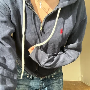 Mörkblå hoodie från Polo Ralph Lauren - Mörkblå hoodie från Polo Ralph Lauren med vit dragsko, dragkedja och klassisk röd logga broderad på bröstet. 