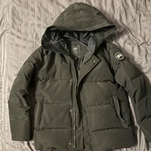 Svart Jacka från Canada Goose M - Säljer en svart dunjacka från Canada Goose i storlek M. Jackan har huva, dragkedja och tryckknappar framtill samt två stora fickor. Klassisk patch på ärmen och quiltad design som håller dig varm under kalla dagar. Black Label