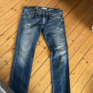 Blå Replay jeans med slitningar - Säljer ett par blå jeans från Replay med snygga slitningar och klassisk femficksdesign. Jeansen har raka ben och en avslappnad passform, perfekt för en cool och avslappnad stil. Materialet är jeans och färgen har en tvättad look.
