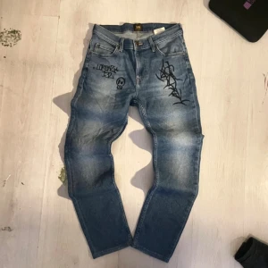 Feta Lee jeans med coola detaljer  - Säljer ett par blå jeans från Lee med unika svarta graffiti- och tribal-inspirerade motiv fram och bak. Jeansen har klassisk femficksdesign, Lee-läderpatch bak och raka ben. Materialet är denim i bomull och passformen är normal.