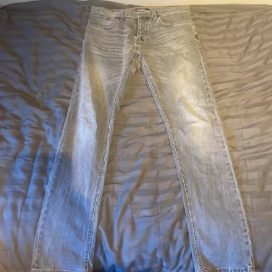 Grå jeans från Nudie Jeans Co - Säljer ett par grå jeans från Nudie Jeans i rak modell. Jeansen är tillverkade i 100% ekologisk bomull och har klassisk femficksdesign med knappgylf. Snyggt slitna detaljer och ljus tvätt för en avslappnad look.