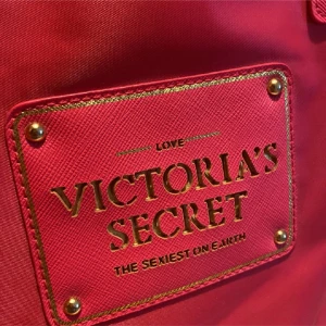 Rosa shoppingväska från Victoria's Secret - Snygg rosa shoppingväska från Victoria's Secret med guldiga detaljer och logga framtill. Väskan har två rejäla handtag och är tillverkad i ett tåligt syntetmaterial. Perfekt för att bära med sig allt man behöver under dagen.