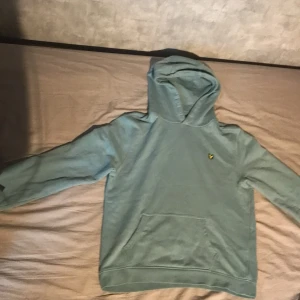 Blå hoodie från Lyle & Scott - Snygg blå hoodie från Lyle & Scott med klassisk gul logga på bröstet. Tröjan har huva, magficka och långa ärmar. Perfekt för chill dagar eller när du vill ha en avslappnad stil.