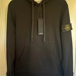 Svart hoodie från Stone Island - Svart hoodie från Stone Island med klassisk logotyp-patch på vänster ärm. Tröjan har huva med breda snören och en stor magficka framtill. Tillverkad i mjuk bomull, perfekt för en avslappnad och stilren look. 