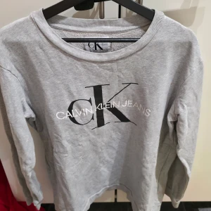 Grå Calvin Klein sweatshirt i bomull - Snygg grå sweatshirt från Calvin Klein Jeans med stor CK-logga framtill. Tröjan är i 100% bomull och har en klassisk rund halsringning samt långa ärmar. Perfekt för en chill och stilren look.