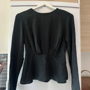 Svart omlottblus med öppen rygg - Säljer en svart blus med lång ärm och snygg omlottdesign i ryggen som ger en öppen rygg-effekt. Blusen har markerad midja och lätt utsvängd nederdel för en feminin siluett. Perfekt för dig som gillar stilrena och trendiga plagg.