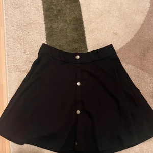 Svart kortkjol med knappar framtill - Svart A-linjeformad kortkjol med snygga silverfärgade knappar längs hela framsidan. Kjolen har en enkel och stilren design som passar till många olika looks. Materialet känns mjukt och följsamt, perfekt för dig som gillar minimalistisk stil.