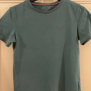 Blå t-shirt från Tommy Hilfiger - Snygg blå t-shirt från Tommy Hilfiger med klassisk rund hals och liten broderad logga på bröstet. T-shirten är i mjuk bomull och har korta ärmar. Perfekt för en clean och stilren look.