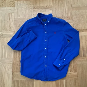 Blå skjorta från Polo Ralph Lauren - Klassisk blå skjorta från Polo Ralph Lauren med lång ärm och knappar framtill. Skjortan har den ikoniska röda broderade loggan på bröstet och är tillverkad i mjuk bomull. Perfekt för en stilren och avslappnad look.