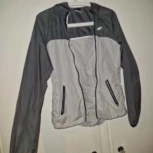 Vindjacka - Nike vindjacka i svart och grått med rutmönster, hel dragkedja och två snedställda fickor med dragkedja. Jackan har huva och reflekterande Nike-logga på bröstet samt reflekterande prickar på ryggen. Perfekt för blåsiga dagar och sportiga outfits. Sparsamt använd fåtal gånger. Ett litet hål på högra sidan nertill, inget som stör