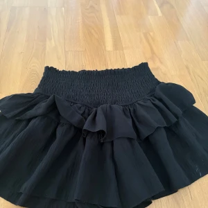 Svart volangkjol från SHEIN - Supersöt svart kortkjol från SHEIN med bred smock i midjan och volangdetaljer upptill. Kjolen har en flowy känsla och är perfekt för dig som gillar en feminin och trendig look. Tillverkad i lätt och luftig polyester.